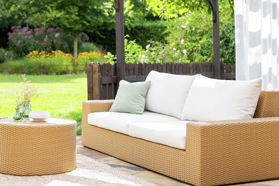 Garden rattan sofa with white cushions and matching rattan pouffe in garden.