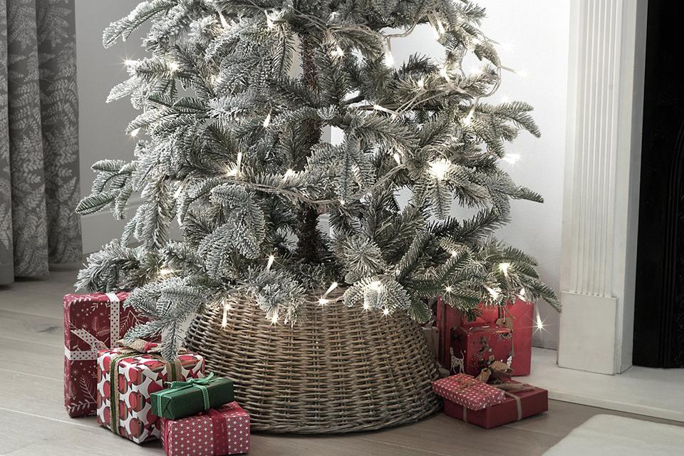 A wicker Christmas tree skirt wrapped around a snowy tree with presents around it.