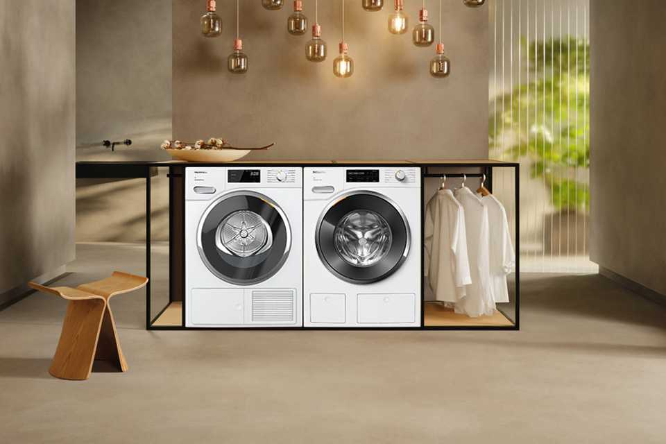 A white Miele washing machine and tumble dryer placed next to each other in an earthy toned room.