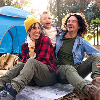 A family of 3 smiling and enjoying outside their tent.