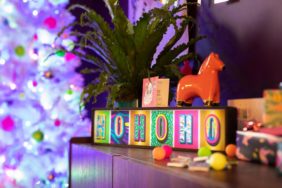 A brightly coloured lit-up sign reading "Ho Ho Ho" on a dresser with a bright Christmas tree behind it.