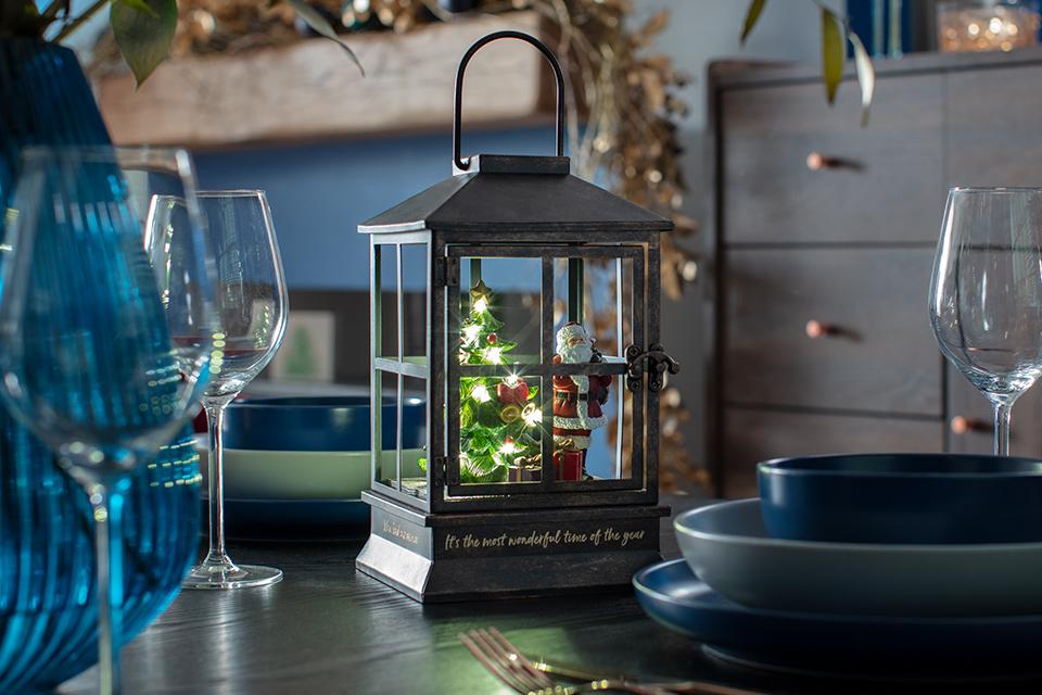 A grey lantern with a lit-up Christmas tree and Father Christmas inside, sat on a dining room table.