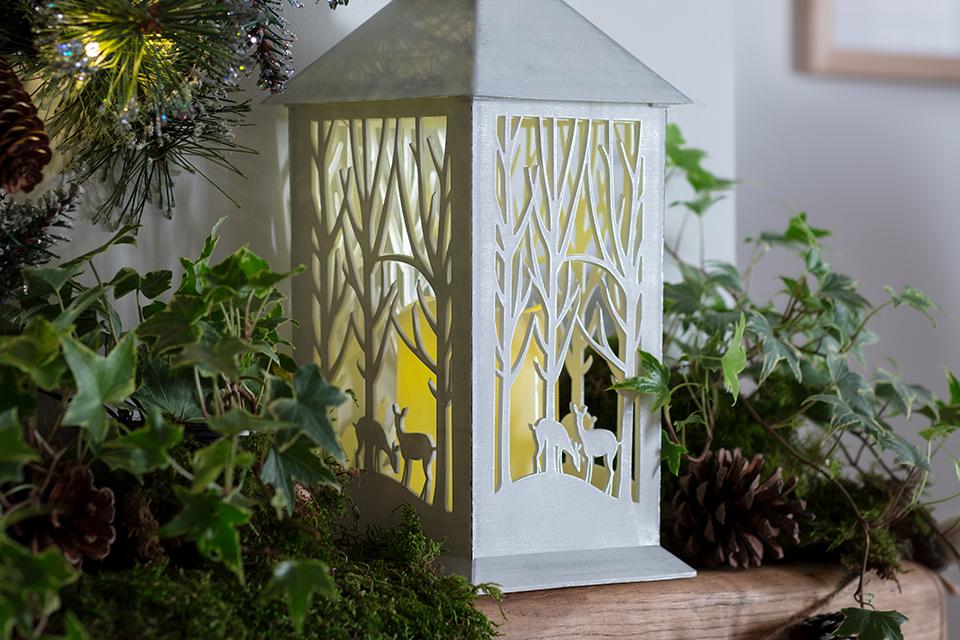 A white lantern with cut out images of deer in a forest, sitting on a mantelpiece.