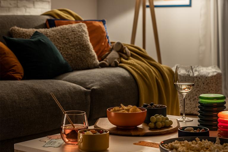 Image of a sofa with lots of cushions and blankets next to a coffee table with lots of snack on it.