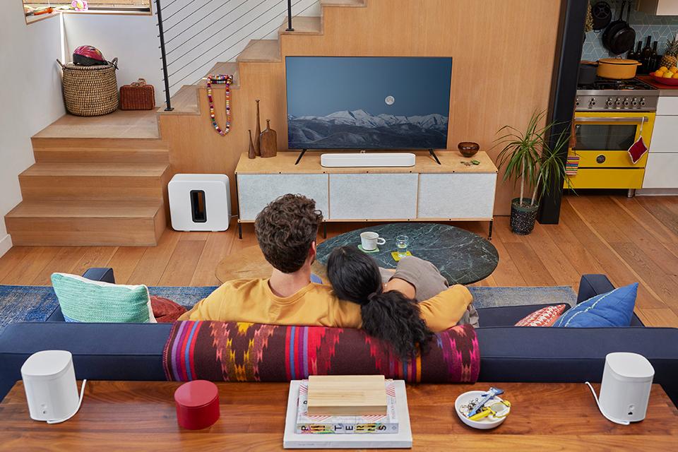 Image shows a couple watching TV, with Sonos speakers arranged around the room for surround sound.
