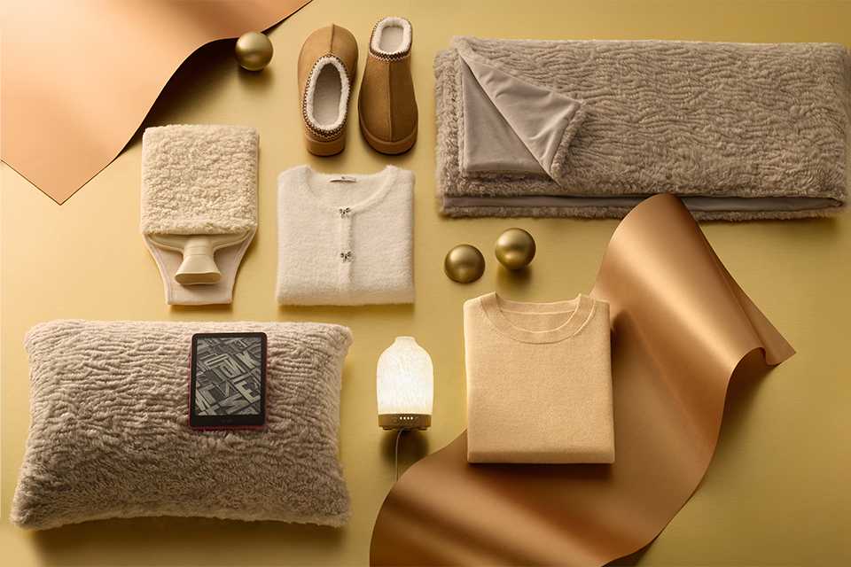 Flat lay of cozy winter essentials in neutral tones, including fluffy beige pillow with e-reader, soft throw blanket, cream cardigan, folded sweater, warm slippers, and decorative gold accents on a golden background.