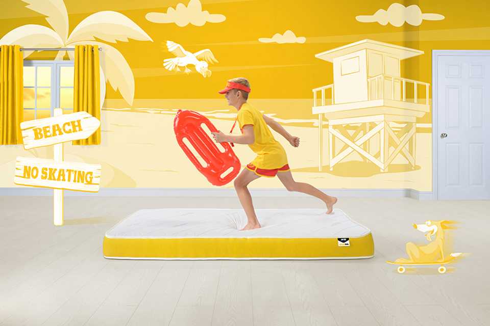 A boy on a Jay-Be® children’s mattress with a surfing board against an animated beach backdrop.