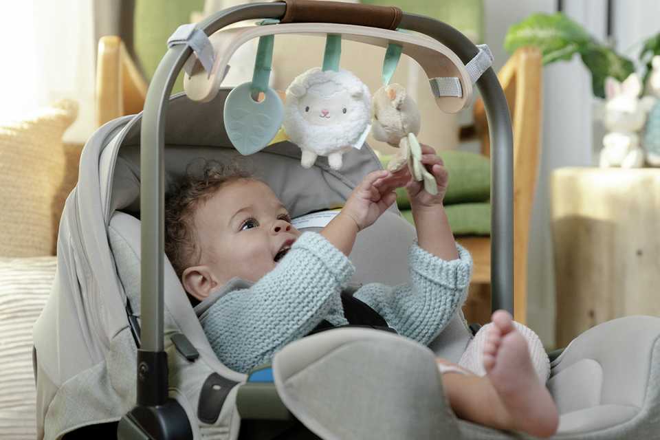 A baby playing with an Ingenuity Wooden Toy Bar attached to a car seat.