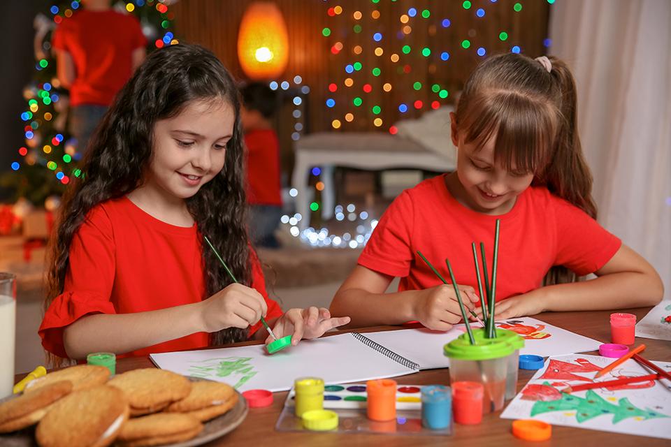 Two young girls sat at dining table painting, with Christmas lights and decorations behind them.