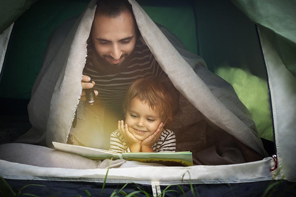 Father and son under blanket in a tent, reading a book together in torchlight.