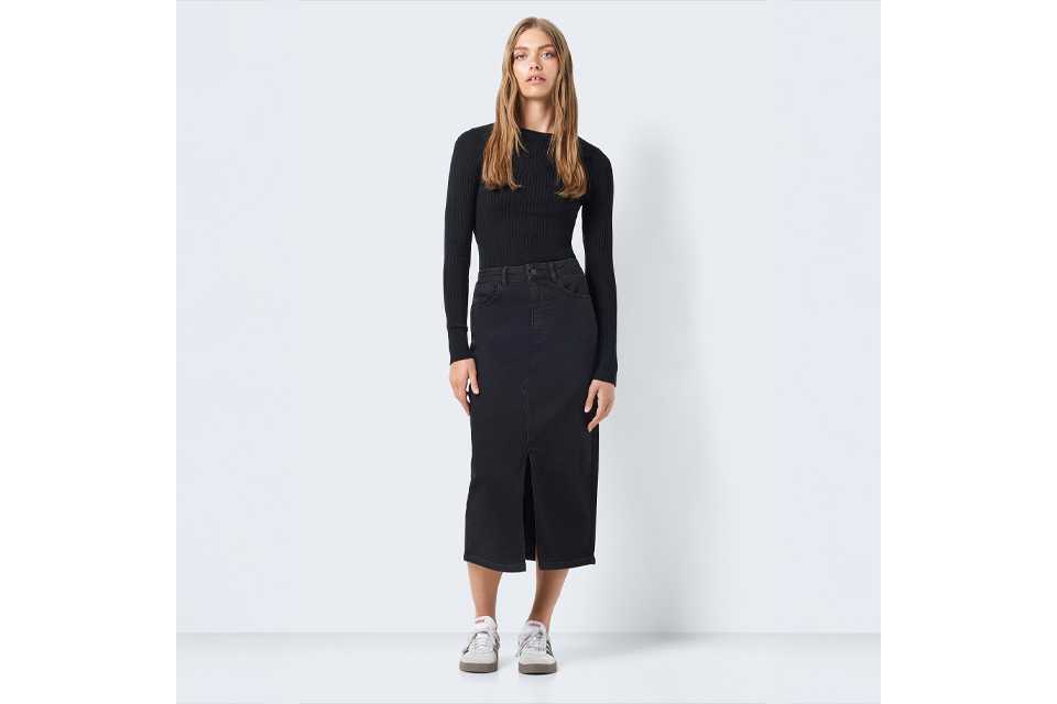 Person wearing a black long-sleeve ribbed top paired with a high-waisted black denim midi skirt featuring a front slit, styled with white sneakers.