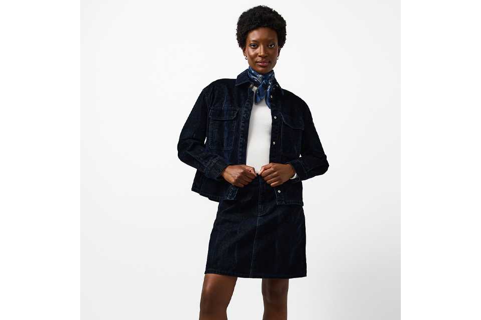 Person wearing a dark denim jacket and matching mini skirt, styled with a white top, blue patterned neck scarf, brown socks, and black loafers.