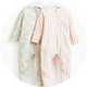Two baby sleepsuits hanging on white hangers, one in pastel green with white heart pattern and the other in pink and white stripes, both featuring zip-up fronts and footed design.