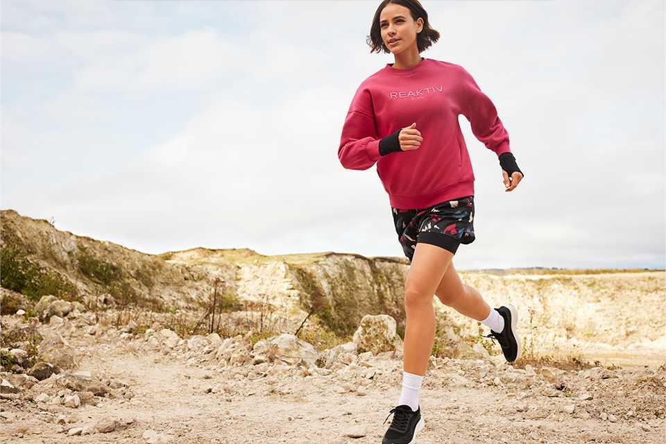 Person running outdoors on a rocky trail, wearing a bright pink sweatshirt with "REAKTIV" text, patterned shorts, white socks, and black running shoes.