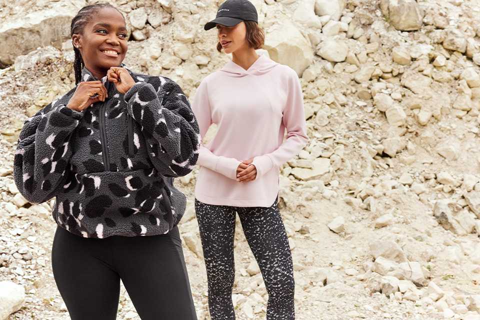 Two people standing on rocky ground wearing activewear; one in a black patterned fleece and leggings, the other in a pink hoodie and speckled leggings.