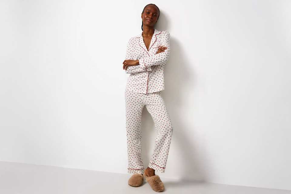 Person standing against a white wall wearing a white pajama set with small black and red heart prints, red piping detail, and beige fluffy slippers.
