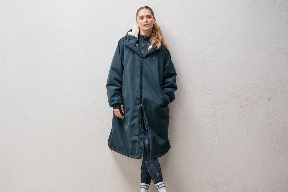 Person standing against a light-colored wall wearing a long dark green hooded coat with a white fleece lining, patterned leggings, white socks, and black athletic shoes.