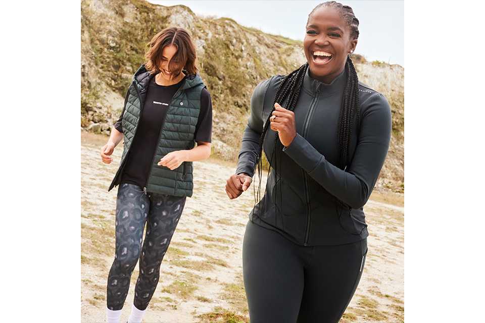 Two people jogging outdoors on rocky terrain. One is wearing a dark zip-up jacket and matching leggings, while the other is in a black T-shirt, patterned leggings, and a quilted vest.
