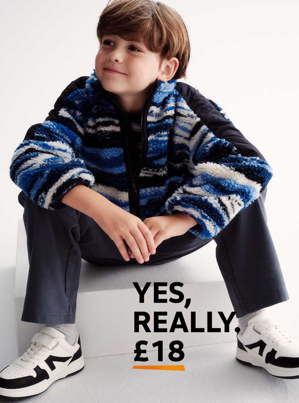 Person sitting on a white block wearing a blue, black, and white patterned zip-up jacket, dark pants, and white sneakers with black accents.