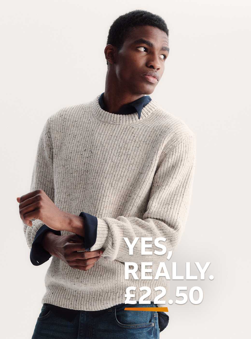 A man wearing a neutral jumper and a dark shirt, with jeans.