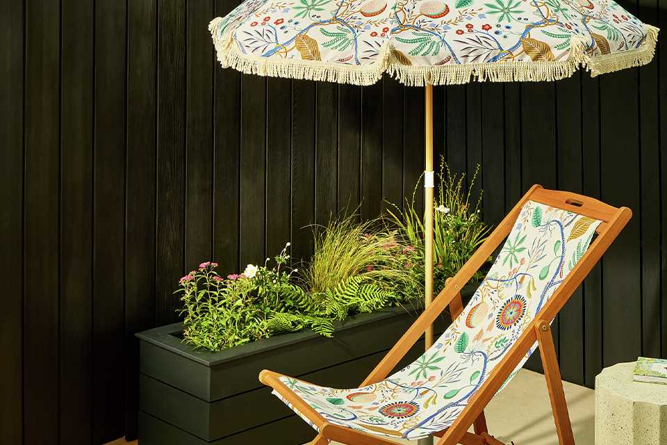 Scion x Habitat garden umbrella and deck chair in white floral pattern next to garden planter with green plants.