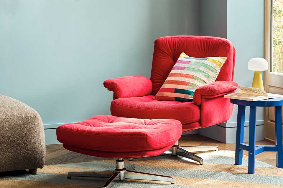 Red velvet accent arm chair with matching pouffe in front and striped multicolour cushion with blue side table next to it and yellow mushroom lamp on top and large round mirror on the wall.