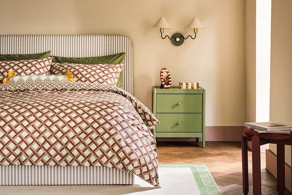 Neutral bedroom with geometric patterned bedding with striped headboard as a vocal point and sage green bed side table with two arm wall light with white shades.