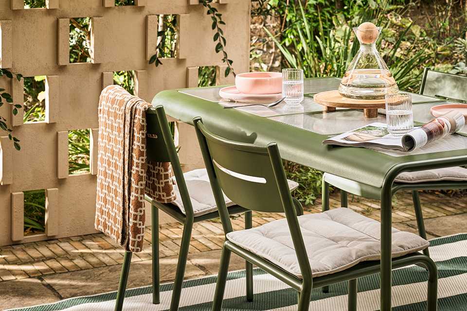 Garden scene showing green table and chairs with striped green and white umbrella for shade and floral seat cushions.