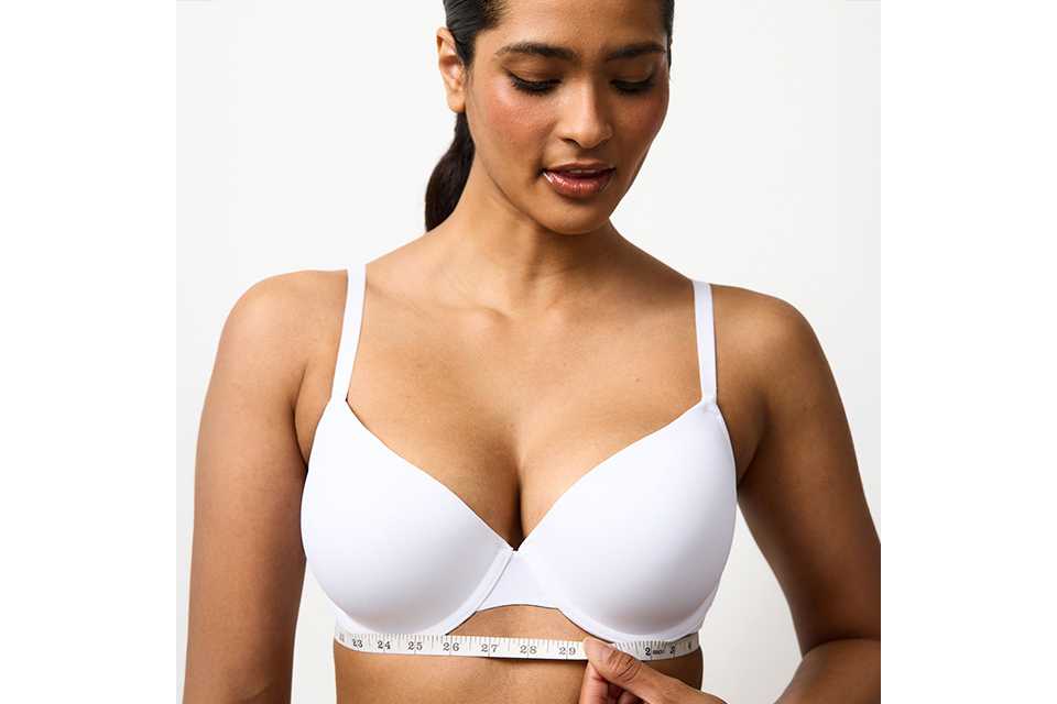 Woman wearing a white bra measuring under her bust using a tape measure.