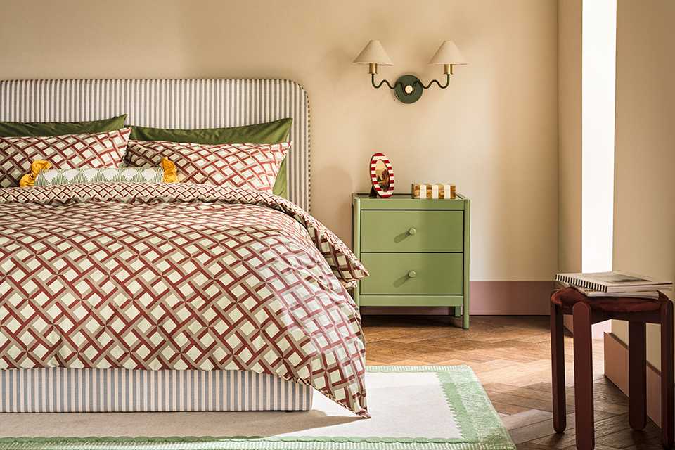 Neutral bedroom with geometric patterned bedding with striped headboard as a vocal point and sage green bed side table with two arm wall light with white shades.