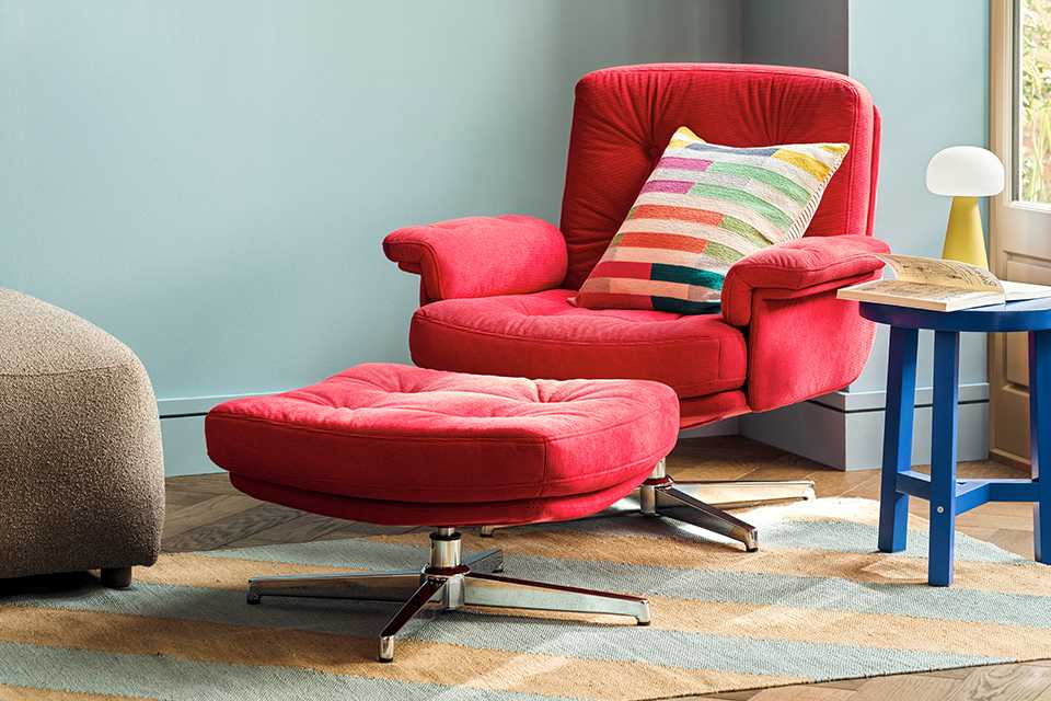 Red velvet accent arm chair with matching pouffe in front and striped multicolour cushion with blue side table next to it and yellow mushroom lamp on top and large round mirror on the wall.