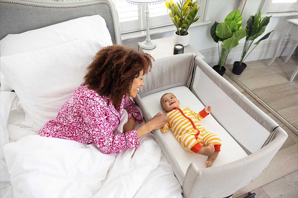 A mother in bed checking on happy baby in a roomie™ go travel bedside crib.