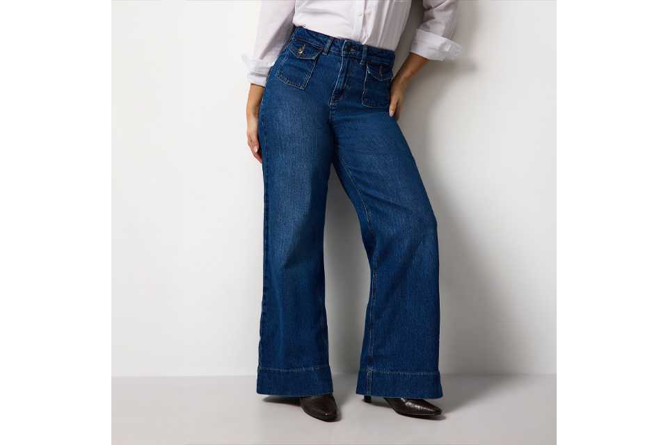 Person standing against a light concrete wall wearing a long-sleeved white lace blouse paired with high-waisted, wide-leg pink trousers and black pointed shoes.