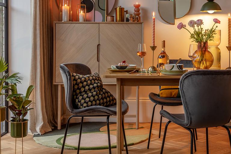 A wooden dining table paired with grey velvet chairs in a dining room displaying tableware, candles and a vase with flowers.