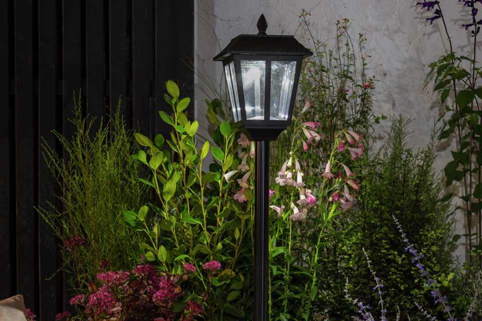 A black Argos Home Metro solar powered lampost in a garden next to some flowers.