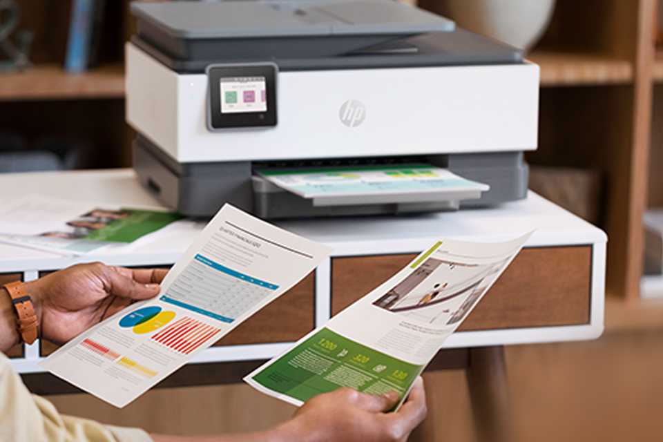 A person holding 2 printed documents in front of an HP office printer.