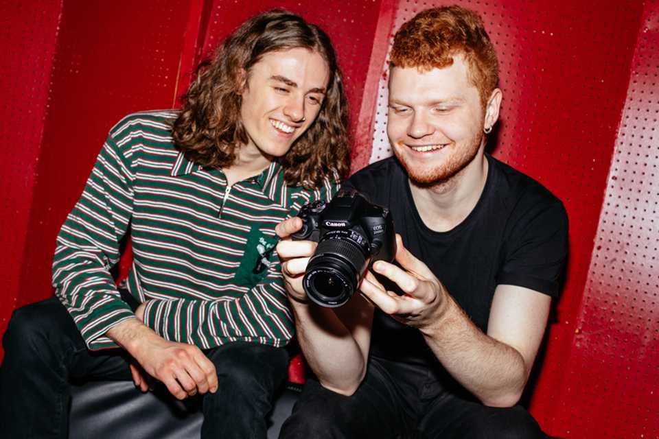 Two friends smiling while looking at pictures on a Canon EOS 2000D camera.