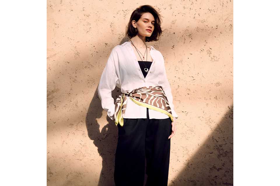 White lightweight shirt styled with black wide‑leg trousers, a patterned waist scarf and neutral woven slip‑on flats in an outdoor setting.