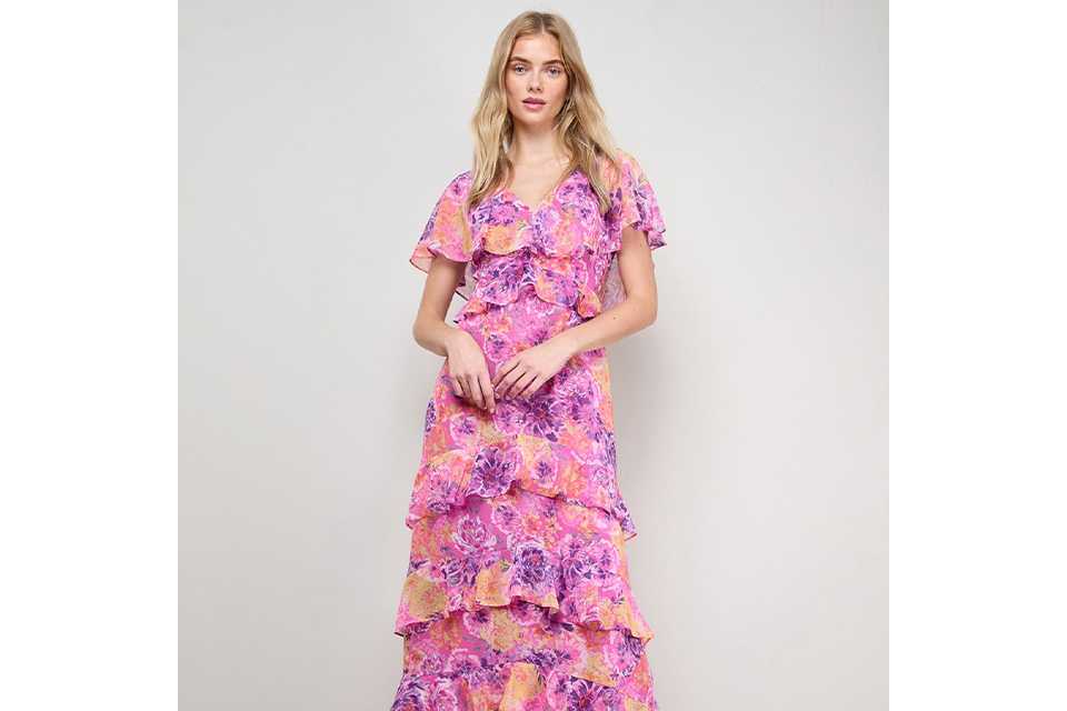 Pink floral tiered midi dress with short flutter sleeves and V‑neckline, styled with neutral heeled sandals on a light studio background.
