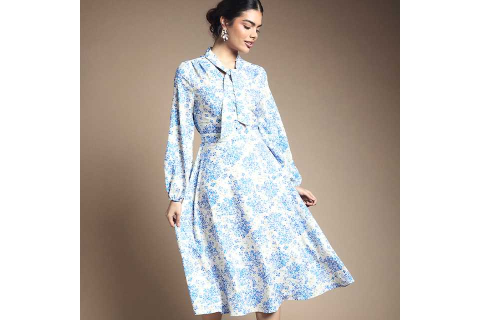 Blue and white floral long‑sleeve midi dress with tie‑neck detail and softly flared skirt on a neutral studio background.