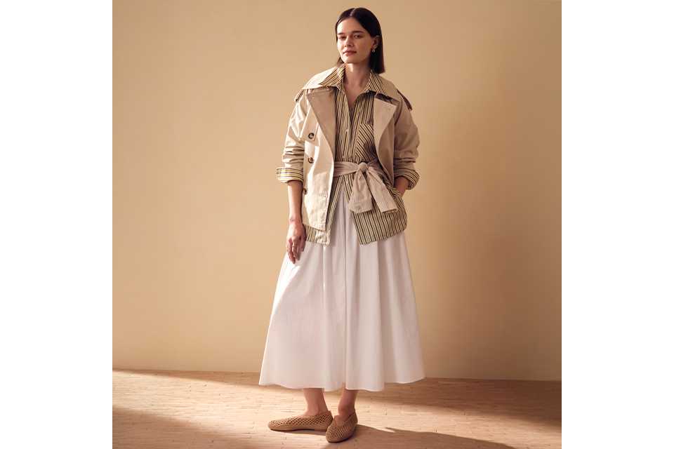 A women wearing a white maxi skirt with a cream striped shirt and cream short trench jacket.