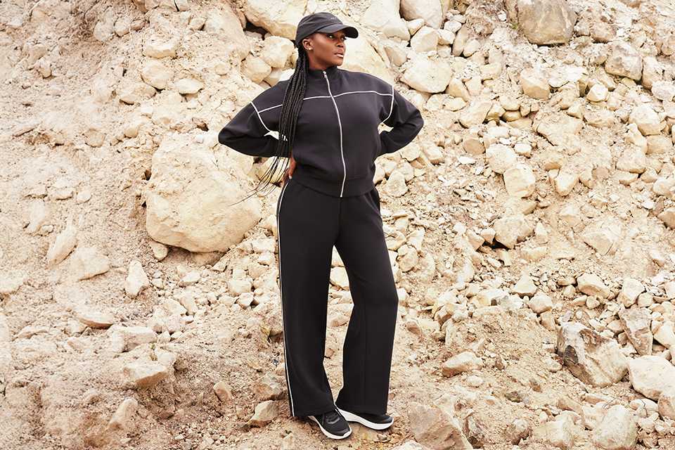 Person standing on rocky terrain wearing a black zip-up jacket with white piping, matching wide-leg pants, black sneakers, and a black cap.