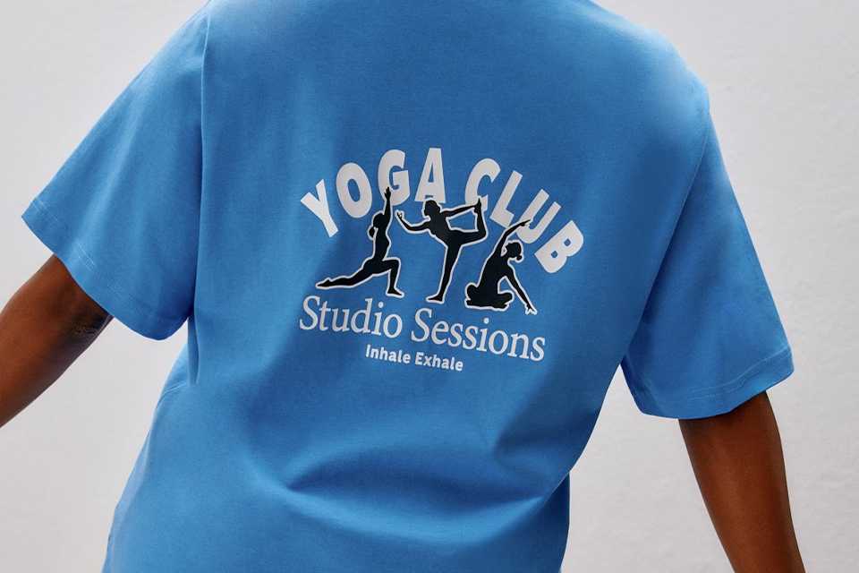 Person lying on a yoga mat, wearing a long-sleeve white top and navy leggings, holding one leg bent, and a person standing in a wide lunge pose on a yoga mat, wearing a long-sleeve blue top and navy pants.