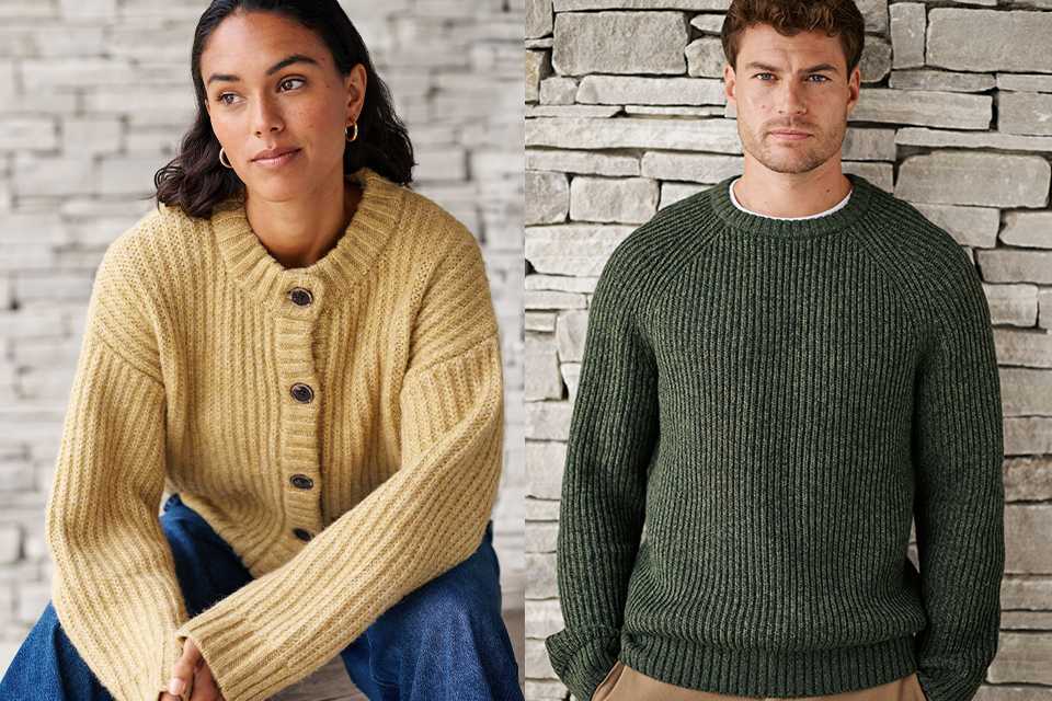 Two people wearing chunky knit sweaters, one in dark green and the other in mustard yellow, seated against a stone wall.