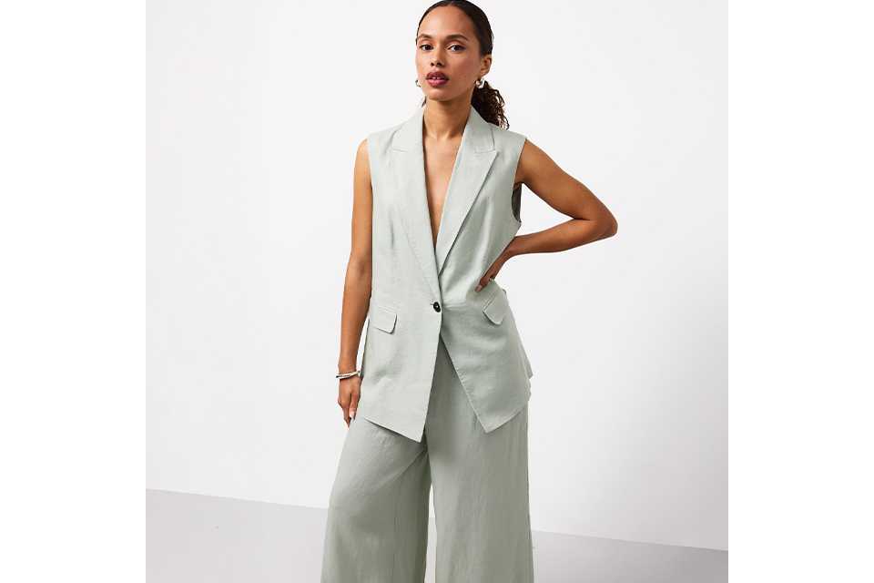 Black tailored blazer and belt styled with a green and blue patterned midi skirt, black heeled sandals and a black shoulder bag on a white background.