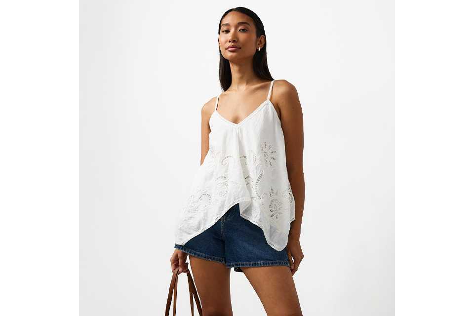 Woman wearing a white cut out cami top, with denim shorts, sandals and a straw bag.