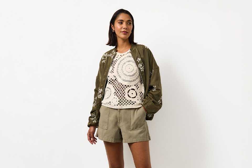 Woman wearing khaki shorts and a crochet top, with an embroidered khaki jacket and sandals.