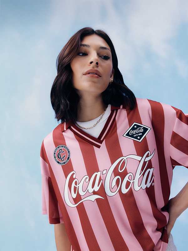 A woman wearing a pink and red stripe football top, with a white top underneath and white skirt, with a gold necklace.