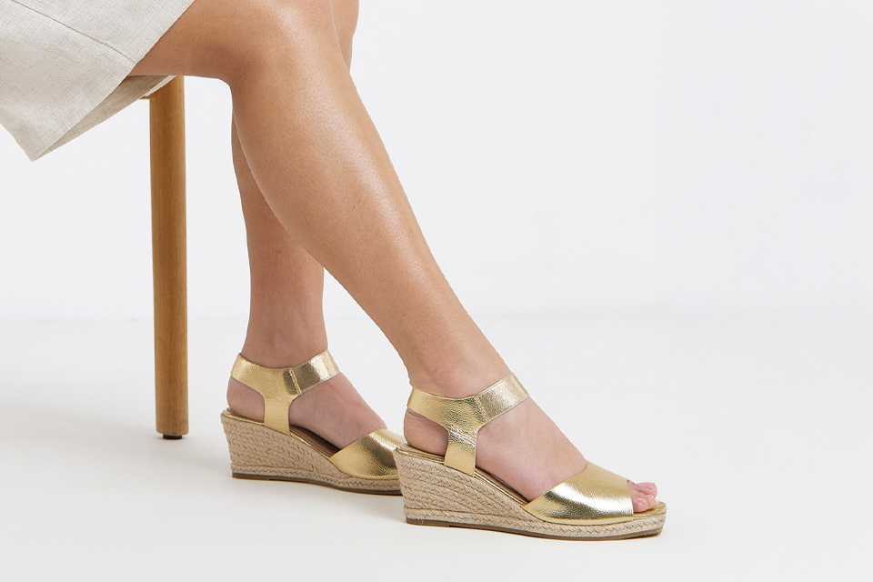Woman sitting down showing her legs, with gold wedge shoes.