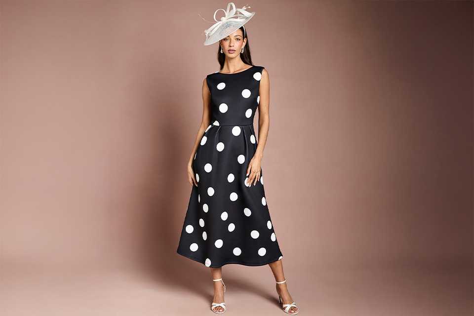 Woman wearing a black dress with white polka dots, a white fascinator and white shoes.